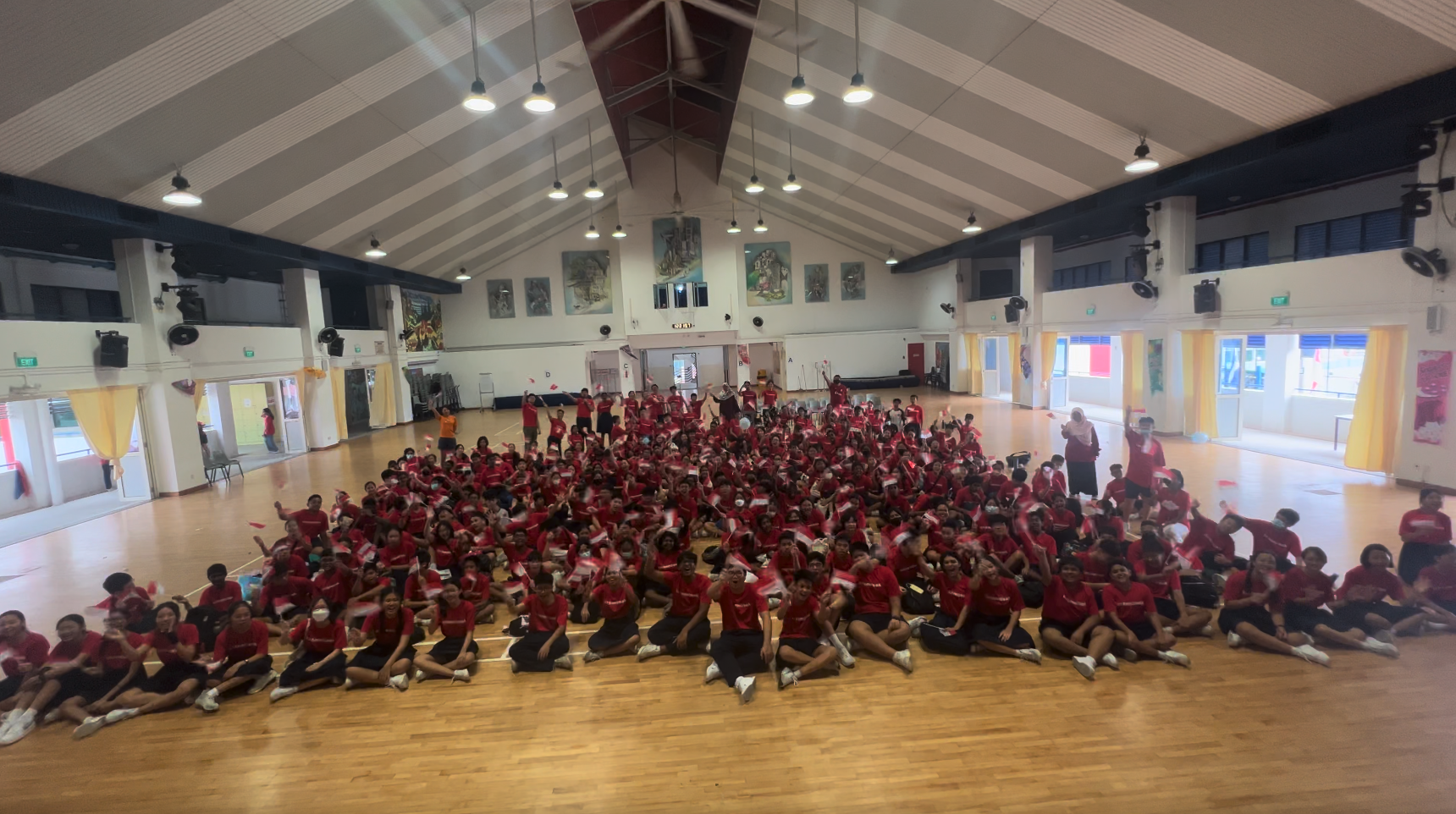 Large group of students in red shirts sitting and posing together on a wooden gymnasium floor.